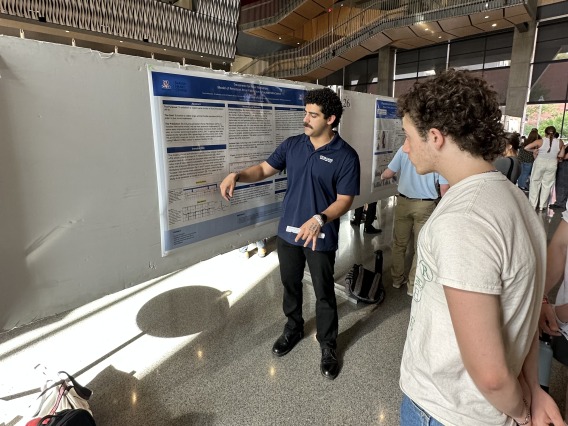 A person wearing a UA polo standing in front of a poster presenting their research.
